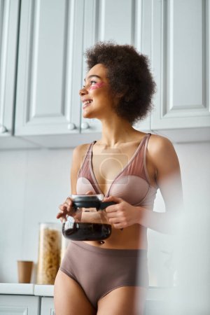 Radieuse jeune femme afro-américaine avec des patchs tenant une cafetière dans une cuisine moderne ensoleillée