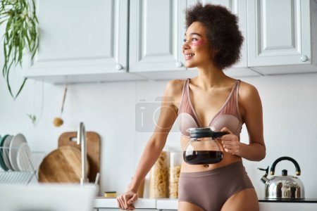 fröhliche junge afrikanisch-amerikanische Frau mit Augenklappen hält Kaffeekanne in einer sonnenbeschienenen modernen Küche
