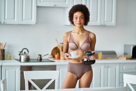 Foto de Radiante joven afroamericana mujer con parches para los ojos sosteniendo taza y cafetera en la cocina - Imagen libre de derechos