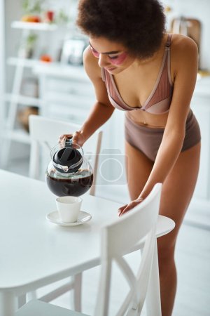 junge und glückliche afrikanisch-amerikanische Frau mit Augenklappen, die in der Küche Kaffee in Porzellantasse gießt