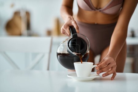 cropped view of african american woman in underwear pouring coffee into porcelain cup in kitchen