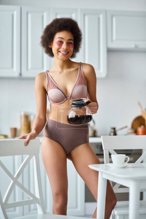 rizado feliz afroamericana mujer con parches para los ojos de pie en ropa interior con cafetera