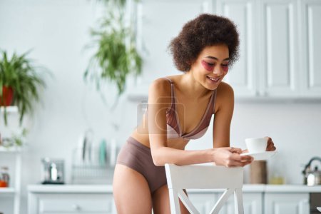 glückliche afrikanisch-amerikanische Frau mit Augenklappen, die in Unterwäsche steht und den morgendlichen Kaffee genießt