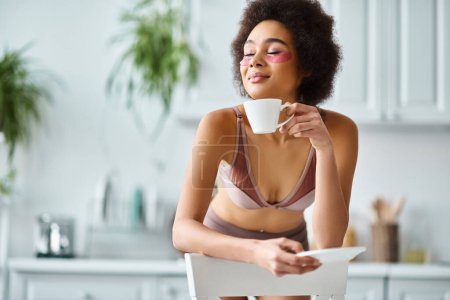 mujer afroamericana complacida con parches para los ojos de pie en ropa interior y disfrutando del café de la mañana