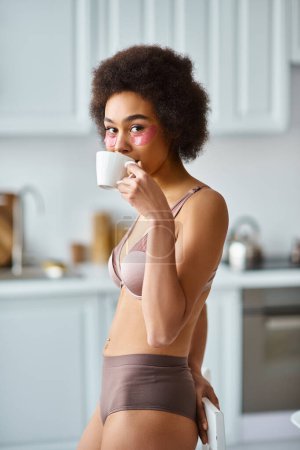 Téléchargez les photos : Heureuse femme afro-américaine avec des patchs pour les yeux debout en sous-vêtements et en buvant le café du matin - en image libre de droit