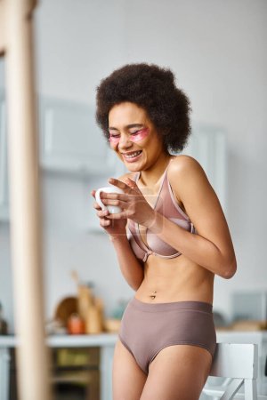 fröhliche afrikanisch-amerikanische Frau mit Augenklappen, die in Unterwäsche steht und Kaffee in der Tasse genießt