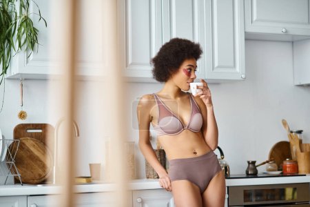 curly african american woman with eye patches standing in underwear and drinking coffee in cup