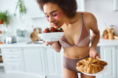 Foto de Mujer negra feliz en lencería disfrutando del olor de las fresas y sosteniendo croissants para el desayuno - Imagen libre de derechos