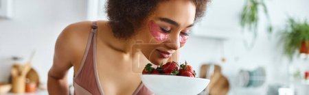 young african american woman in lingerie smelling fresh strawberries in kitchen, banner