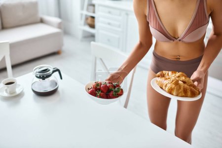 cropped african american woman in lingerie holding plates with croissants and strawberries