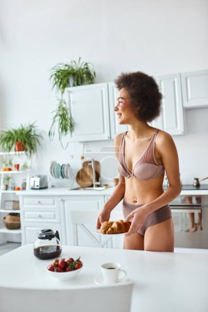 happy african american woman in lingerie serving croissants and strawberries for breakfast