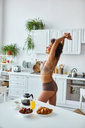 smiling african american woman in lingerie stretching near delicious breakfast on kitchen table
