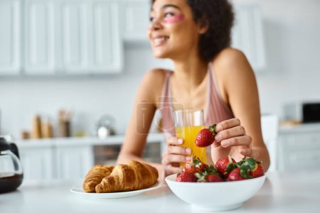 happy african american woman holding fresh strawberry and orange juice during breakfast at home