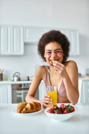 happy african american woman eating fresh strawberry and holding glass of orange juice in kitchen