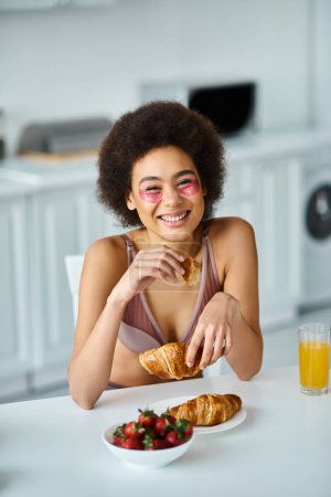 Téléchargez les photos : Joyeuse femme afro-américaine tenant croissant fraîchement cuit pendant le petit déjeuner dans la cuisine - en image libre de droit