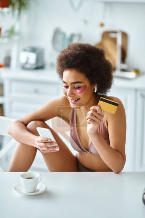 Lächelnde Afroamerikanerin kauft online mit Kreditkarte und Smartphone in der Küche ein