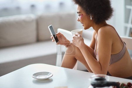 Smiling african american woman in lingerie with eye patches enjoying coffee and browsing smartphone
