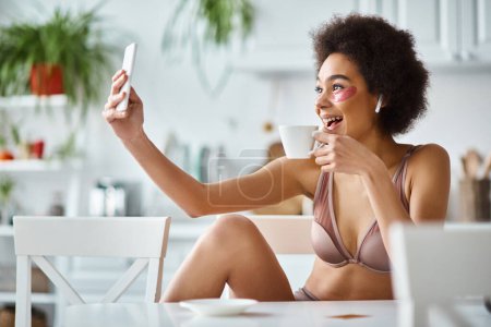 Smiling african american woman in lingerie with eye patches taking selfie with cup of coffee