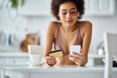 Téléchargez les photos : Femme afro-américaine gaie faisant du shopping en ligne avec carte de crédit et smartphone dans la cuisine - en image libre de droit