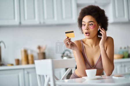 überraschte Afroamerikanerin mit Kopfhörern und Flecken unter den Augen beim Betrachten ihrer Kreditkarte