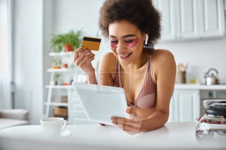 Téléchargez les photos : Joyeuse femme afro-américaine faisant du shopping en ligne avec tablette avec carte de crédit à la main et café de côté - en image libre de droit