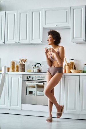 Serene moment of happy african american woman in earphones and lingerie sipping morning coffee