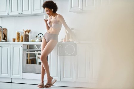 Serene moment of joyful african american woman in earphones and lingerie sipping morning coffee