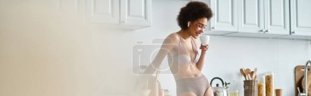 Téléchargez les photos : Bannière de femme afro-américaine heureuse en écouteurs et lingerie sirotant le café du matin dans la cuisine - en image libre de droit