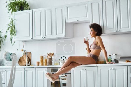 cheerful african american woman in lingerie holding cup of coffee and sitting on kitchen worktop