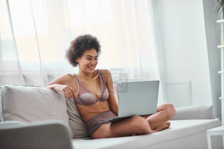 Foto de Mujer afroamericana feliz en lencería sentada en el sofá y viendo la película de comedia en el ordenador portátil - Imagen libre de derechos