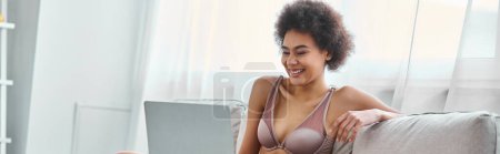 mujer afroamericana feliz en lencería sentada en el sofá y viendo la película en el ordenador portátil, pancarta