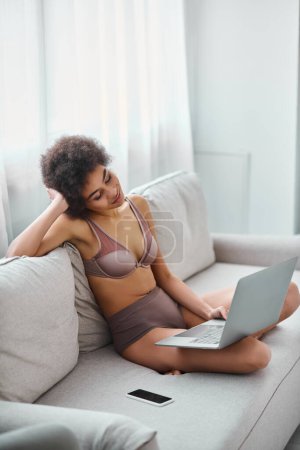 Téléchargez les photos : Heureuse femme afro-américaine en lingerie assis sur le canapé et regarder des films sur ordinateur portable à la maison - en image libre de droit