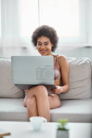 happy african american woman in lingerie sitting on couch and watching funny movie on laptop at home