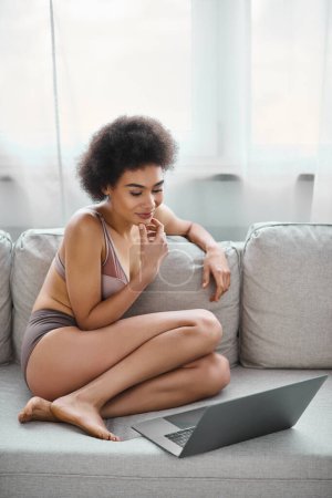 pensive african american in lingerie working remotely on laptop while sitting on cozy sofa at home