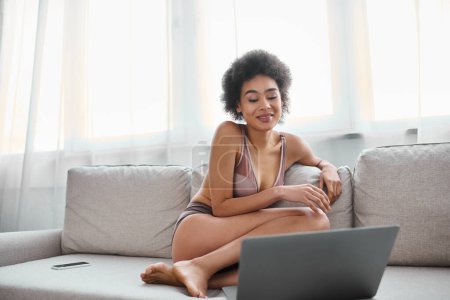 Foto de Mujer afroamericana en lencería sentada en el sofá y sonriendo mientras ve la película en el portátil - Imagen libre de derechos