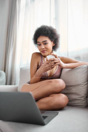 african american woman in lingerie sitting on sofa and holding cup while watching movie on laptop