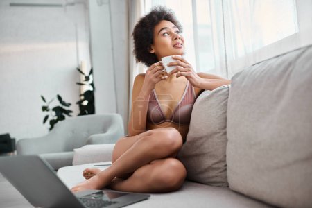 Foto de Mujer afroamericana soñadora en lencería sosteniendo la taza mientras está sentado en el sofá cerca de la computadora portátil, mirando hacia arriba - Imagen libre de derechos