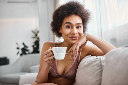 mujer afroamericana feliz en lencería sosteniendo taza de café y mirando a la cámara