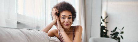 retrato de mujer afroamericana joven y rizada en lencería mirando a la cámara en casa, pancarta