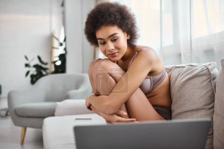 african american woman in lingerie sitting on sofa and watching movie on laptop, weekend vibes