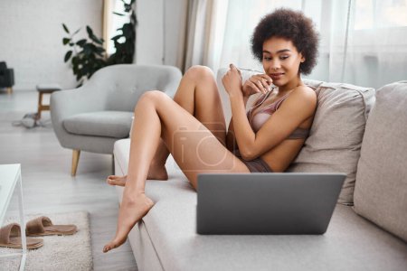 mujer afroamericana rizada en lencería sentada en el sofá y viendo películas en el portátil, vibraciones de fin de semana