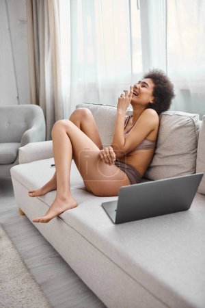 Téléchargez les photos : Africaine américaine femme en lingerie assis sur canapé et rire de film comique sur ordinateur portable, profiter - en image libre de droit