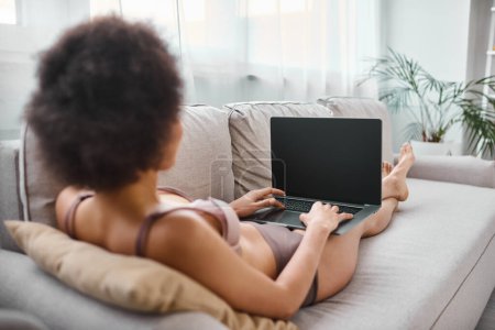 back view of african american in lingerie working remotely on laptop while sitting cozy sofa at home