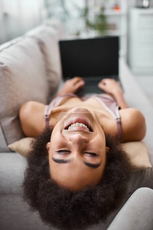 positiver Afroamerikaner in Dessous, der ferngesteuert am Laptop arbeitet, während er zu Hause auf dem gemütlichen Sofa sitzt