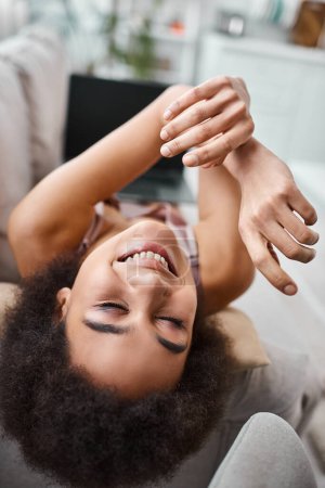 Foto de Mujer afroamericana positiva en lencería descansando en el sofá con el ordenador portátil borroso en sus vueltas - Imagen libre de derechos