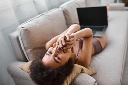 Afroamerikanerin in Dessous sitzt auf Sofa und lacht aus Comedy-Film auf Laptop, Spaß