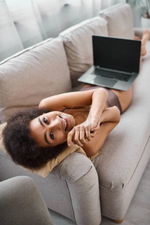 glückliche afrikanisch-amerikanische Frau in Dessous auf dem Sofa liegend mit Laptop auf dem Schoß, am Wochenende