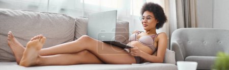 bannière de femme afro-américaine en lingerie et lunettes de travail à distance sur ordinateur portable dans le salon
