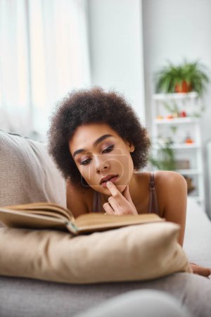 Konzentrierte junge Afroamerikanerin liest ein Buch in Dessous auf einem bequemen Sofa