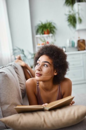 mujer afroamericana soñadora y joven leyendo un libro en lencería y acostada en un cómodo sofá